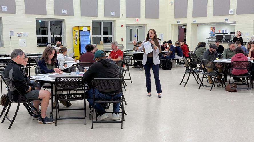 A woman leads a discussion in a school cafeteria as adults sit at tables reviewing papers and participating in small-group conversations.