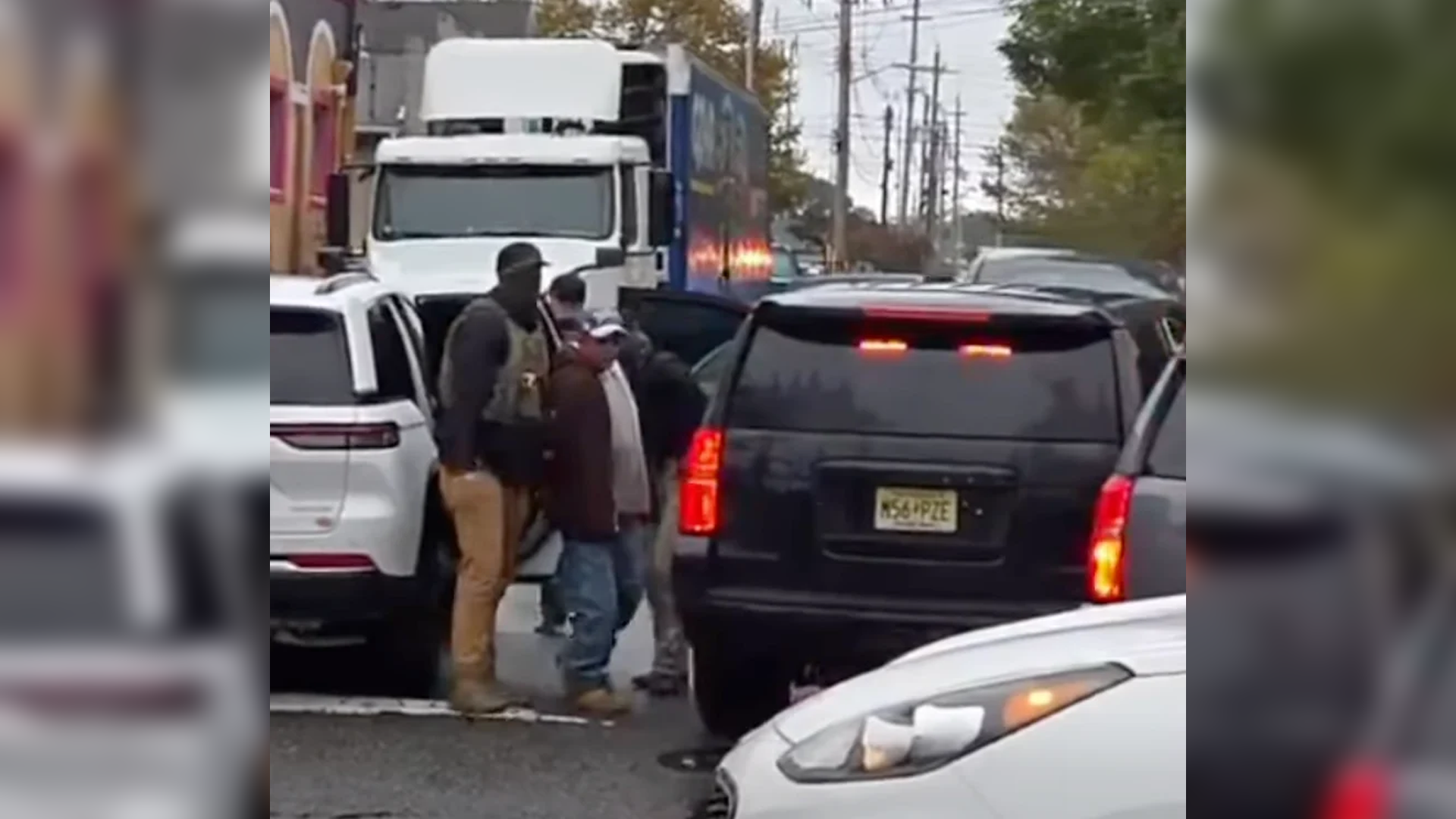 Federal immigration agents escort a man between vehicles on a street in Red Bank, New Jersey, surrounded by traffic.