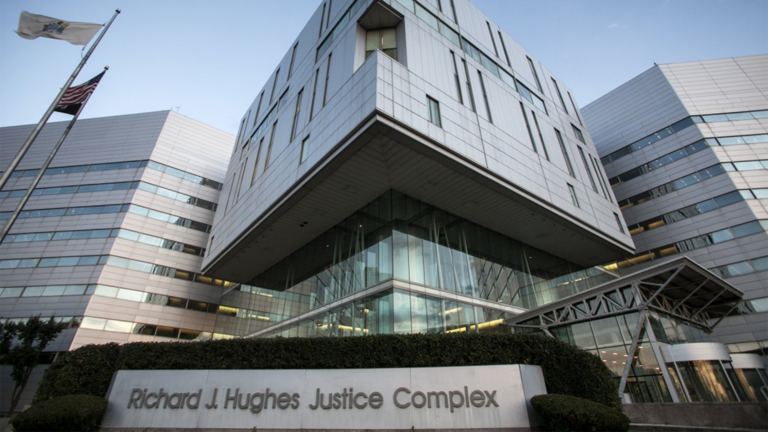 Exterior of the Richard J. Hughes Justice Complex in Trenton, New Jersey, featuring a modern, multi-story building with glass windows, silver paneling and the building’s name displayed on a stone sign in front. An American flag and a New Jersey state flag fly outside.